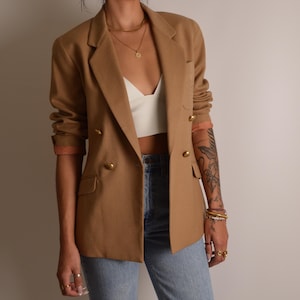 K&ouml;nnte beinhalten: Ein beigefarbener zweireihiger Blazer mit goldfarbenen Kn&ouml;pfen, fallendem Revers und einer Brusttasche. Der Blazer wird &uuml;ber einem wei&szlig;en bauchfreien Oberteil und einer hellblauen Jeans getragen, wobei die &Auml;rmel pfirsichfarben gef&uuml;ttert sind.