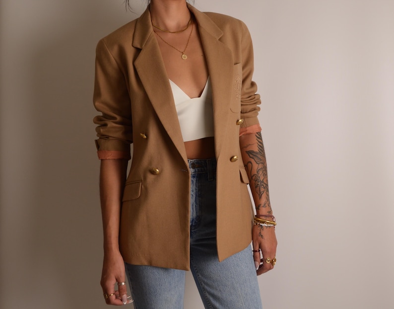 K&ouml;nnte beinhalten: Ein beigefarbener zweireihiger Blazer mit goldfarbenen Kn&ouml;pfen, fallendem Revers und einer Brusttasche. Der Blazer wird &uuml;ber einem wei&szlig;en bauchfreien Oberteil und einer hellblauen Jeans getragen, wobei die &Auml;rmel pfirsichfarben gef&uuml;ttert sind.