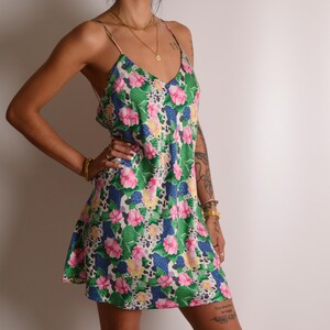 May include: A short slip dress featuring a vibrant floral pattern with pink flowers, green leaves, and blue accents on a white background. The dress has thin spaghetti straps, a V-neckline, and a relaxed, straight-cut silhouette that falls above the knee.