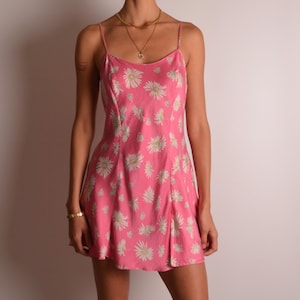 May include: A bright pink mini dress featuring a repeating white daisy floral pattern, thin spaghetti straps, and a scoop neckline. The dress has a straight hemline and a fitted silhouette that falls above the knee, showcasing a simple design.