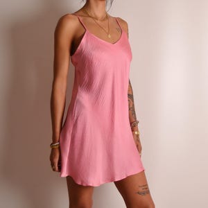 May include: A short, sleeveless pink slip dress featuring a V-neckline and thin spaghetti straps. The fabric has a subtle, crinkled, textured finish throughout, and the garment falls to a hemline above the knee with a loose, flowing silhouette.