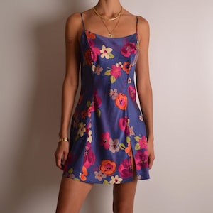 May include: A short, sleeveless blue dress featuring a vibrant floral pattern in shades of pink, orange, and cream. The garment has thin spaghetti straps, a straight neckline, and a small vertical slit on the lower left side of the hemline.