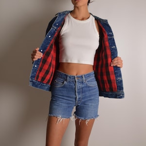 May include: A denim jacket with a red and black plaid lining is worn open over a white tank top. The jacket and shorts are blue. The shorts have a frayed hem.
