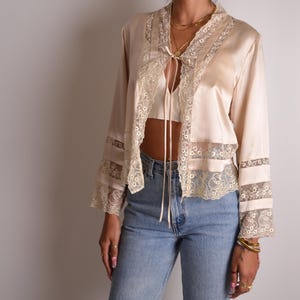 Könnte beinhalten: Eine langärmelige Jacke aus champagnerfarbenem Satin mit cremefarbener Spitzenverzierung an der Vorderseite, am Saum und an den Ärmelbündchen. Die Jacke verfügt über einen dünnen Satin-Bindegürtel am Ausschnitt und eine hellblaue Jeans.