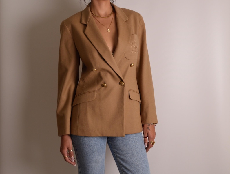 K&ouml;nnte beinhalten: Ein beigefarbener zweireihiger Blazer mit fallendem Revers, langen &Auml;rmeln und goldfarbenen Kn&ouml;pfen. Die Jacke verf&uuml;gt &uuml;ber zwei Pattentaschen an der Taille und eine Brusttasche mit gesticktem Logo, kombiniert mit einer hellblauen Jeans.