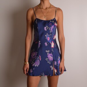 May include: A navy blue mini dress featuring a satin-like finish and a vibrant floral pattern in shades of purple, pink, and white. The dress has thin spaghetti straps, a straight neckline, and a vertical slit on the lower left side of the hem.