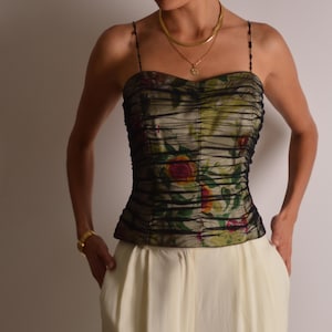 Könnte beinhalten: Ein Bustier-Top mit floralem Print und Spaghettiträgern. Das Oberteil hat ein gerafftes Design mit einer Mischung aus grünen, roten und gelben Blumenmustern. Das Model trägt eine goldene Halskette und ein Armband sowie eine cremefarbene Hose.