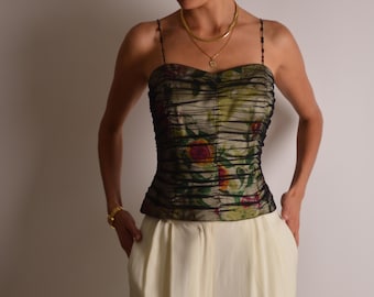 May include: A floral print bustier top with spaghetti straps. The top features a ruched design with a mix of green, red, and yellow floral patterns. The model is wearing a gold necklace and bracelet, and cream-colored pants.