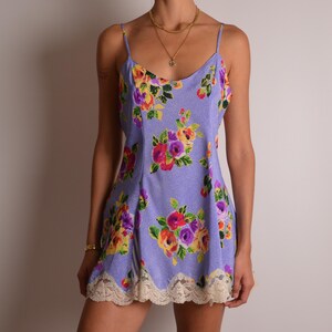 May include: A light purple slip dress featuring a small white polka dot pattern and a vibrant floral print with red, yellow, and purple roses. The garment has thin spaghetti straps, a scoop neckline, and a scalloped cream lace trim at the hem.