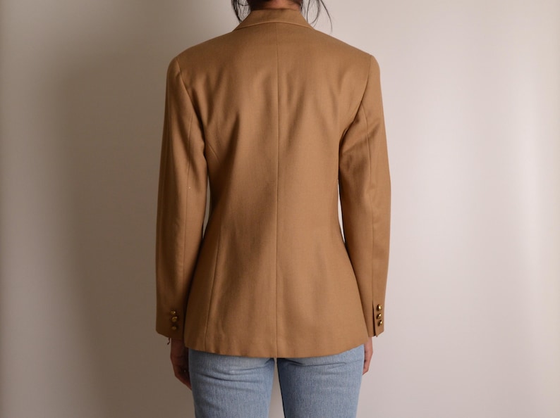 K&ouml;nnte beinhalten: Ein beigefarbener Blazer mit Kragen, langen &Auml;rmeln und drei kleinen goldfarbenen Kn&ouml;pfen an jedem &Auml;rmelabschluss. Das Kleidungsst&uuml;ck ist von hinten zu sehen und zeigt eine taillierte Passform mit vertikalen N&auml;hten sowie eine hellblaue Jeans.