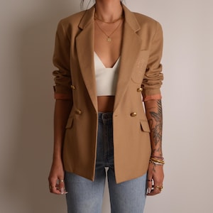 K&ouml;nnte beinhalten: Ein beigefarbener zweireihiger Blazer mit goldfarbenen Kn&ouml;pfen, fallendem Revers und zwei Pattentaschen auf der Vorderseite. Die &Auml;rmel sind hochgeschoben und geben den Blick auf ein pfirsichfarbenes Futter frei. Dazu ein wei&szlig;es bauchfreies Top.