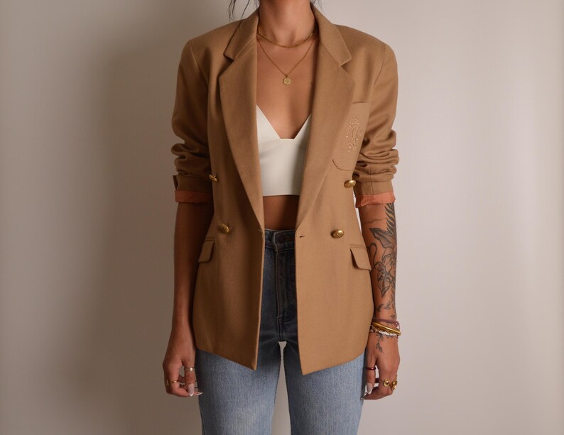 K&ouml;nnte beinhalten: Ein beigefarbener zweireihiger Blazer mit goldfarbenen Kn&ouml;pfen, fallendem Revers und zwei Pattentaschen auf der Vorderseite. Die &Auml;rmel sind hochgeschoben und geben den Blick auf ein pfirsichfarbenes Futter frei. Dazu ein wei&szlig;es bauchfreies Top.