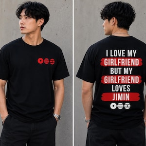 May include: A black short-sleeve t-shirt featuring a small red graphic on the front chest. The back displays the white text "I LOVE MY GIRLFRIEND BUT MY GIRLFRIEND LOVES JIMIN" over three red brushstroke bars, with three small white circular icons below.