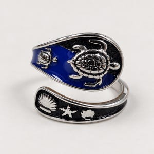 Puede incluir: Un anillo abierto de tono plateado con una sección de esmalte azul que presenta dos tortugas marinas en relieve y una sección de esmalte negro con una concha, una estrella de mar y una concha más pequeña en relieve. Es ajustable.