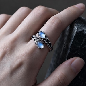 May include: An open-style silver ring featuring two teardrop-shaped moonstones. The metal band displays intricate filigree detailing and a twisted rope texture surrounding each stone, creating a balanced and symmetrical appearance on the finger.