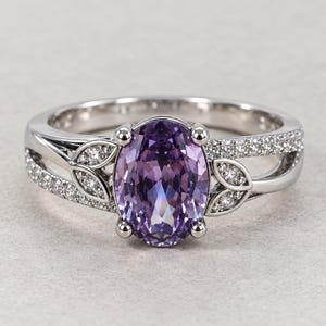 May include: A silver-toned ring featuring a large, oval-cut purple gemstone at the center. The band is split, adorned with small, clear pavé-set stones and decorative metal leaf-shaped accents flanking the central purple stone on both sides.