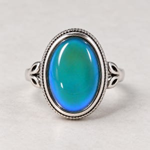 May include: A silver-toned ring featuring a large, oval-shaped mood stone that displays a gradient of teal, green, and blue colors. The setting is framed by a beaded border, and the band includes decorative cut-out leaf-like metal details.