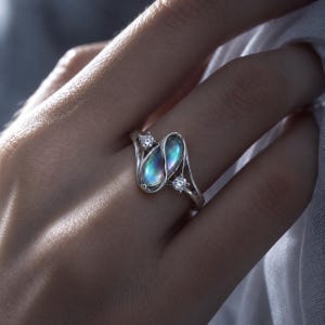 May include: A silver ring featuring a central oval-shaped iridescent moonstone-like gem with a blue glow, flanked by two small round clear stones. The metal band has a curved, flowing design that wraps around the central gemstone setting.