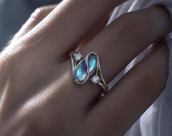 Könnte beinhalten: Ein silberner Ring mit einem ovalen, irisierenden Stein mit blauem Schimmer in der Mitte, flankiert von zwei kleinen runden, klaren Steinen. Die Metallschiene hat ein geschwungenes, fließendes Design, das die zentrale Fassung umschließt.