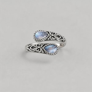 May include: A silver bypass ring featuring two teardrop-shaped moonstones set in ornate, oxidised metalwork. The band displays intricate scroll patterns and rope-style borders surrounding each iridescent stone, creating a continuous, open-ended design.