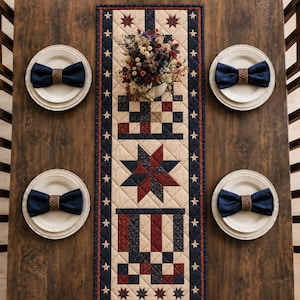 May include: A quilted table runner featuring a red, white, and blue star and block pattern with a border of white stars on a navy background. Four white plates are set with navy blue napkins held by woven rings, and a floral centerpiece sits atop.