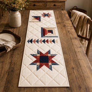May include: A rectangular quilted table runner featuring a cream-colored background with a diamond-stitched pattern. The runner displays a series of patchwork designs including a large eight-pointed star, a row of triangles, and two square blocks.