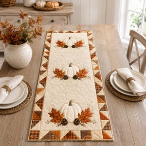 May include: A rectangular quilted table runner featuring a cream-colored center with three embroidered white pumpkins, brown autumn leaves, and pinecones. The border consists of a geometric pattern in shades of orange, brown, and tan fabric.