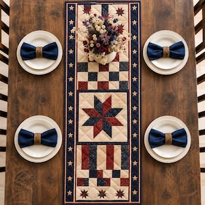 May include: A rectangular quilted table runner featuring a red, white, and blue star and checkerboard pattern, bordered by a navy blue strip with white stars. Four white plates with navy blue napkins held by woven rings are placed around it.