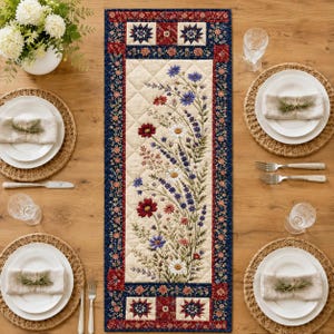 May include: A rectangular quilted table runner featuring a central cream panel with a printed wildflower bouquet of red, blue, and white flowers. The border is navy blue with a red floral pattern and star motifs at the top and bottom ends.