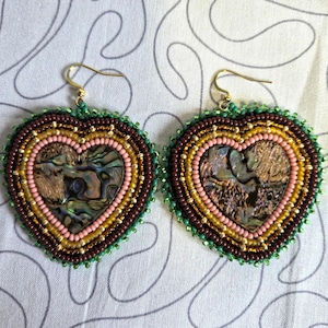 Puede incluir: Un par de pendientes en forma de corazón con una incrustación central de concha de abulón iridiscente rodeada por filas concéntricas de pequeñas cuentas en tonos marrón oscuro, dorado, rosa claro y un borde exterior verde vibrante.