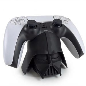 Könnte beinhalten: Ein schwarz-weißer Videospiel-Controller auf einem schwarzen Ständer in Form eines Helms mit dreieckiger Gesichtsmaske und vertikalem Gitter. Der Controller verfügt über zwei Analogsticks, ein Steuerkreuz und vier Aktionstasten.