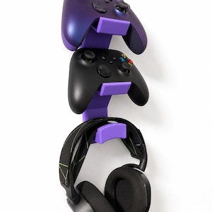 Könnte beinhalten: Eine vertikale violette Wandhalterung mit zwei Gaming-Controllern und einem schwarzen Over-Ear-Kopfhörer. Der obere Controller ist violett schimmernd, der mittlere Controller ist schwarz mit farbigen Tasten und der Kopfhörer hat ein gemustertes Band.