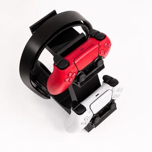 Può includere: Un supporto per accessori da gioco in plastica nera con un arco superiore curvo per cuffie e due slot inferiori per controller. Un controller wireless rosso e un controller wireless bianco sono montati sul supporto in verticale.