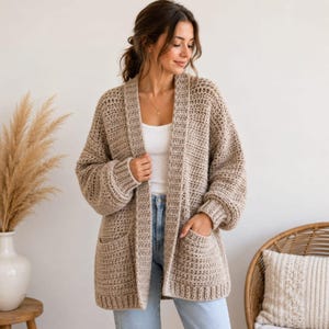 May include: A beige open-front cardigan featuring a chunky crochet knit texture, long sleeves, and two large patch pockets on the front. The garment is worn over a simple white top and blue denim jeans, showcasing a relaxed, loose-fitting silhouette.