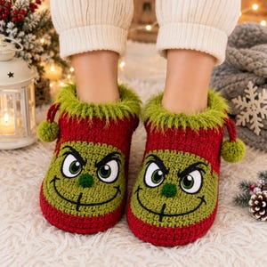 May include: A pair of red and green crochet slippers featuring a grumpy character face with large green eyes, a stitched mouth, and a small green nose. The slippers have a fuzzy green trim at the top and two small green pom-poms on each side.