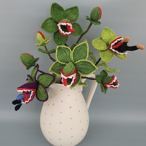 Op de afbeelding: Een crèmekleurige keramieken kan met kleine blauwe stippen bevat een boeket gehaakte vleesetende planten. De groene, paarse en rode garenbloemen hebben open monden met witte tanden en rode of blauwe tongen in verschillende stadia.