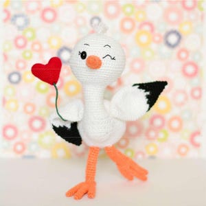 Puede incluir: Un juguete de pájaro tejido a ganchillo en color blanco con cabeza redonda, pico naranja y patas naranjas. El pájaro tiene las puntas de las alas negras y guiña un ojo. Sostiene un pequeño corazón rojo brillante tejido a un tallo verde.