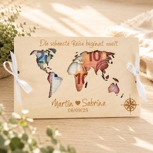 Könnte beinhalten: Eine rechteckige, helle Holzplatte mit einer lasergeschnittenen Weltkarte. Der Text "Die schönste Reise beginnt zweit" ist oben eingraviert, darunter stehen "Martin ♥ Sabrina" und "08/09/23". An den Seiten sind weiße Schleifen befestigt.