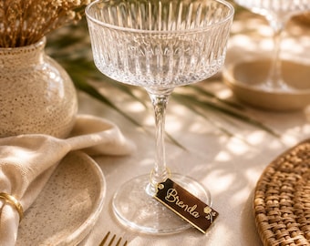 May include: A clear glass coupe with vertical ribbed detailing on the bowl and a slender stem. A small, rectangular, dark-colored acrylic tag is attached to the base with a gold ring, featuring the name Brenda in gold script and a small rose icon.