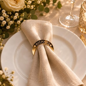 May include: A gold-coloured metal napkin ring featuring a curved design with the name RICHARD engraved in the centre, flanked by two small heart symbols on either side, holding a folded beige linen napkin on a white ceramic dinner plate.