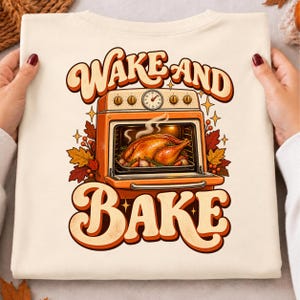 Puede incluir: Una camiseta de color crema con un gráfico de un horno naranja que contiene un pavo asado. El texto "WAKE AND BAKE" está impreso en letras grandes de color naranja de estilo retro sobre y debajo del horno, rodeado de hojas de otoño.