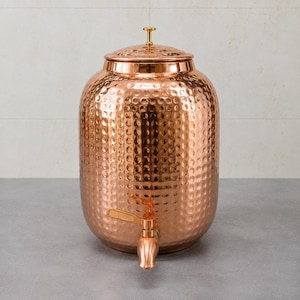 May include: A large, cylindrical copper beverage dispenser featuring a distinct hammered texture across its exterior surface. The vessel includes a matching copper lid with a small gold-toned knob handle and a functional spigot at the base.