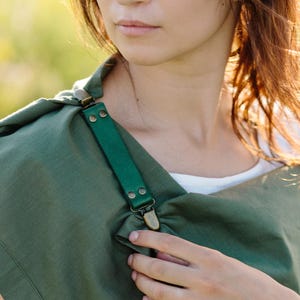 Puede incluir: Una prenda envolvente de color verde oliva con un borde ancho con dobladillo y una correa de cuero verde oscuro asegurada con clips y remaches de tono bronce metálico. La tela tiene una textura tejida visible y se coloca sobre los hombros.