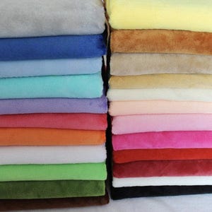 May include: Two vertical stacks of folded fabric squares in a wide variety of solid colours. The left stack features cool tones like grey, blue, teal, and green, while the right stack displays warm tones including yellow, tan, pink, red, and black.