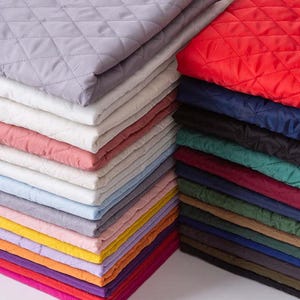 May include: A stack of folded quilted fabric in various colours including light grey, white, pink, light blue, yellow, orange, red, dark blue, black, green, and brown. Each piece features a consistent diamond-shaped stitched pattern throughout.