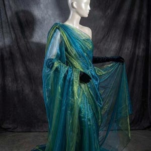 May include: A floor-length, one-shoulder gown crafted from sheer, iridescent fabric that shifts between shades of teal, blue, and lime green. The garment features voluminous, draped layers of translucent material and is paired with long, black velvet gloves.