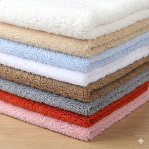 May include: A stack of folded sherpa fleece fabric squares in various colours including white, beige, light blue, brown, grey, orange, and pink. Each square features a thick, textured, and fuzzy pile surface with a consistent soft appearance.