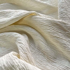 Puede incluir: Una pieza de tela color blanco roto que presenta una superficie texturizada y arrugada con pliegues y ondulaciones profundas. El material muestra un color crema claro uniforme, con sombras que acentúan el patrón fino, lineal y ondulado.