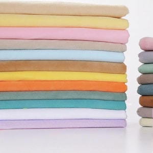 Könnte beinhalten: Ein großer Stapel gefalteter Stoffstücke in verschiedenen Unifarben wie Beige, Gelb, Rosa, Hellblau, Orange, Türkis, Weiß und Lavendel, neben einem kleineren vertikalen Stapel gefalteter Stoffe in Grau-, Grün- und Brauntönen.