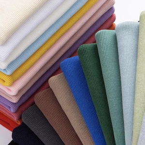 May include: A collection of folded ribbed fabric swatches in a wide variety of colors including white, cream, yellow, pink, purple, red, brown, black, blue, and multiple shades of green, arranged in a layered, fan-like display of textile samples.